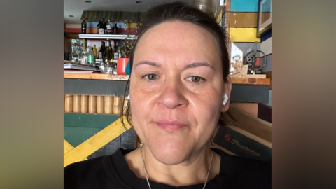 A selfie showing Jodi's head and shoulders. She has a slight smile and has white wireless buds in her ears. She has dark hair. A drinks bar appears to be behind her.