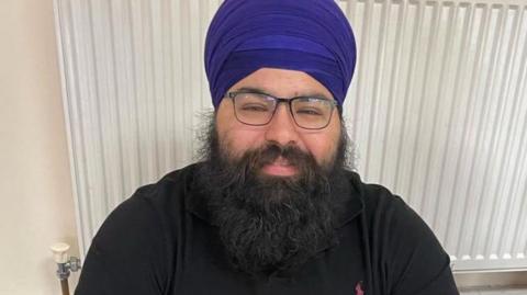 Kuljit Singh Uppal dressed in a black top and blue turban and black glasses. He is sitting on the floor in front of a radiator