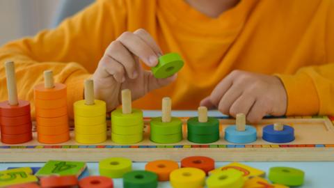 A child is wearing an orange top and is playing with a wooden maths game. The game consists of wooden pegs and coloured pieces to help count. 