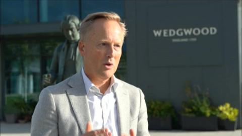 A man with swept back fair hair is wearing a light grey jacket and open-neck white shirt. He is standing outside the Wedgwood building during a TV interview. The building has a modern grey frontage with Wedgwood written on the front in white capitals. On the forecourt is a bronze statue of the company founder, and planters in a row along the front wall.