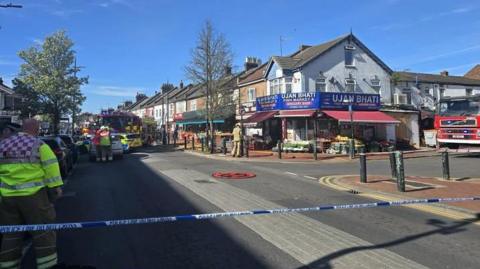 A row of shops with flats above them. The shops are behind some police tape. There are fire engines and firefighters in the road.