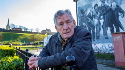 Denis Bradley leaning over a railings looking off camera. He is wearing a coat and scarf. In the background a mural depicting a scene from bloody Sunday.