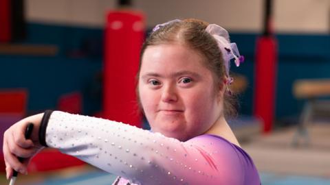 A young woman in a gymnastic training area. She is looking at the camera as she twirls a stick with a ribbon attached to it. She wears a purple and pink leotard. She has a matching coloured scrunchie in her brown hair