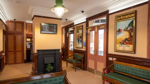 Early 20th Century-inspired waiting room at Castle Cary station, with destination posters of Devon and Cornwall on the walls. Varnished wooden benches with green leather seats and back rests line the walls and there is a fireplace in the centre of the room.