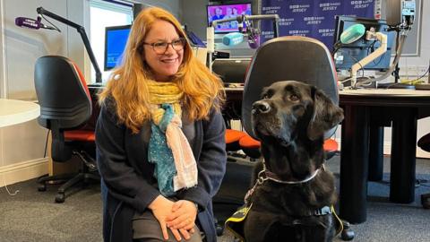 Radio presenter Alison Moss wearing a navy cardigan with a blue, white and yellow scarf is seen next to the retired guide dog who is a black Labrador. 