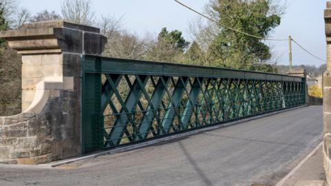 The bridge has a fresh layer of grey tarmac and is bordered by a green, criss-cross metal wall. There are tall stone brick pillars on either side. With tall green trees in the distance.