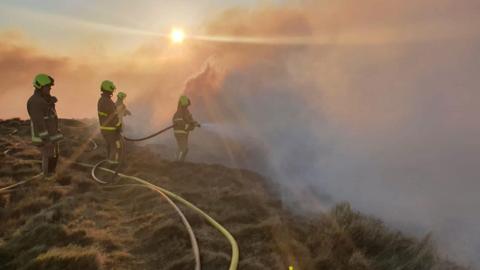 Four fire fighters in a field are holding hoses into thick clouds of smoke, with sunshine beaming through.