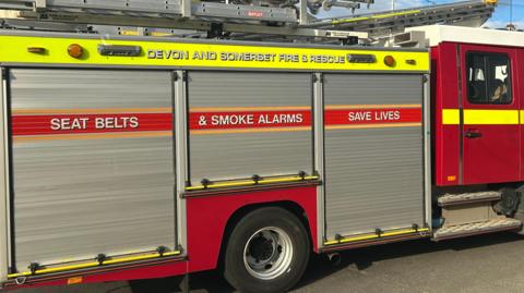 A Devon and Somerset Fire Service engine at the scene of an incident. The engine is red and has large silver shutter doors along the side. It has the service's name written along a yellow strip and has "seat belts and smoke alarms save lives" also written on a red strip along the shutters.