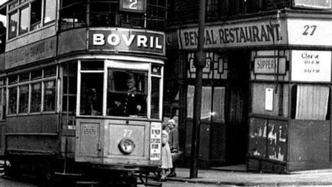 A black and white image of Bradford in the 1940s. A tram is going uphill and behind it is a restaurant that says "Bengal Restaurant" above the door.