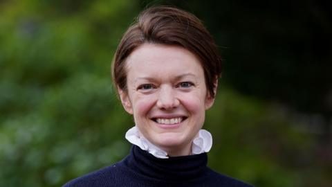 A woman with short, brown hair in a blue roll neck jumper with pink ruffled shirt showing from underneath it