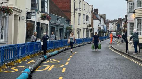 A blue pipe running along a road with shops either side and across a bus stop. Blue barriers are along the pavement on the left-hand side, and pedestrians, some carrying shopping are walking along the road. Workers in hi-vis outfits are in the background 