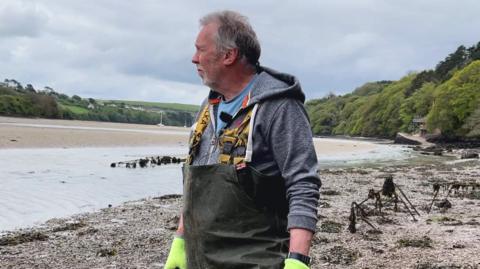 Tim Edwards is wearing waterproof waders, a hoodie, and bright gloves stands on a muddy shore beside shallow water, looking across an estuary. Around them are low racks or frames covered in seaweed, suggesting a shellfish or coastal farming area backed by green hills and trees.