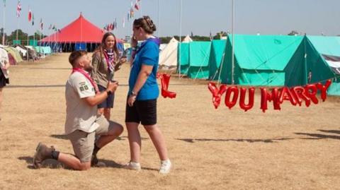 Chris is kneeling on one knee with Hannah covering her mouth and wearing a blue top. 