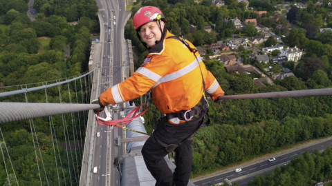 Sam wearing a red hard hat, an orange hi-visibility jacket, black trousers and safety gear stands on a thick cable at the top of a tall suspension bridge, high above a wooded area with green trees, roadways and houses. He is clipped to a harness and looking back at the camera.