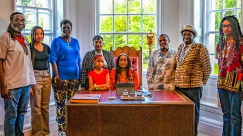 Nine people, all black, dressed in bright colours standing around a desk where a Sally Kimondo is sat. On the desk is a large book and a pen and an iPad. In the background are windows and there are trees beyond.