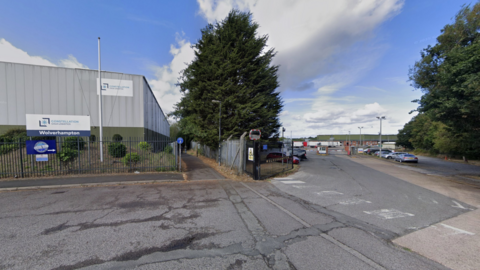 A tarmac road leads to the entrance to the warehouse. A large, grey unit stands on the left with trees standing either side of a driveway entrance.