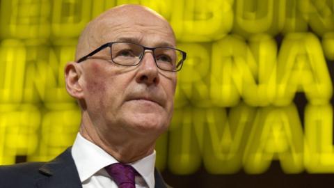 John Swinney, who is bald and wearing glasses, in front of a yellow luminous sign. He is wearing a dark suit, white jacket and purple tie. 