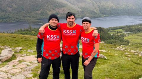 Three men in read tops with KSSX written on them standing on top of a mountain with a lake in the distance.