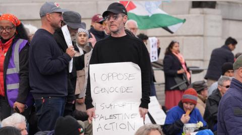 Massive Attack frontman Robert Del Naja standing at a protest at Tralfalgar Square holding a sign that reads: "I oppose genocide I support Palestine Action." He is wearing a black baseball cap, sunglasses and a black jumper