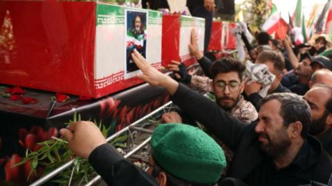 The image depicts a solemn funeral ceremony in Iran. Several red coffins, each draped with the national flag of Iran, are prominently displayed. The flag’s green, white, and red tricolor with Persian script is visible, emphasizing the national significance of the event. One of the coffins bears a portrait of a man in military uniform—likely honouring someone viewed as a martyr or key figure. Mourners are gathered closely, some reaching out to touch the coffins, while others hold green sprigs