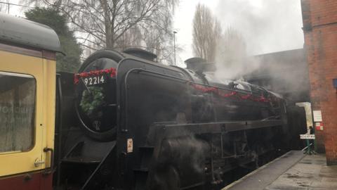 A black steam locomotive is at a Great Central Railway station in Leicestershire. It has red tinsel draped along the side of it and there is a green wreath on the front of the vehicle.