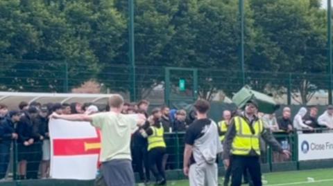 Football fans stood on a pitch. Some are stood behind a gate. There is a bin in the air. There are trees in the background. 