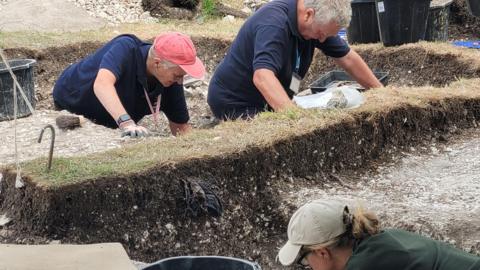 Volunteers wearing blue t-shirts stand in trenches at the Highdown archaeological dig.