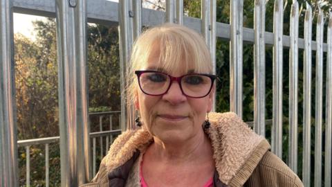 Teri Trickett looking down the camera, she's wearing glasses and a brown jacket with a fluffy collar. Her blonde hair is tied back. Behind her you can see the metal fences with spikes at the top, and beyond that the shrubbery and trees of the Glebe field are visible. 