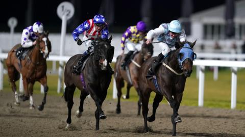 Two jockeys on horses, and two behind them, are racing on a sandy track at Chelmsford City Racecourse at night-time. The white fencing is visible and a building in the background out of focus.