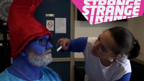 A woman painting the beard of a man dressed as a smurf and the Strange logo