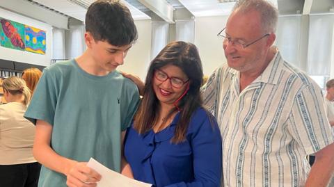 Ethan is wearing a teal T-shirt and he is holding a white piece of paper. His mother and father are looking on pleased.