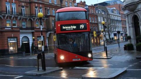 A modern red London bus turns a corner at a pedestrian crossing in central London. The signage on the front reads Battersea Bridge 19.