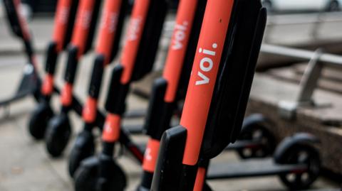 A neat row of red Voi electric scooters parked on a pavement. Only the one in the foreground is in focus, while the red are blurry. 