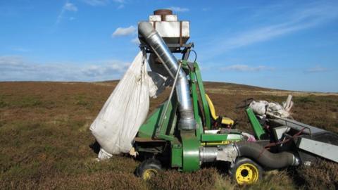 A piece of green machinery with a white sack at one end, held up by a green metal arm on wheels