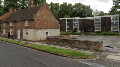 Two contrasting buildings face an empty asphalt road: In the foreground on the left behind a strip of grass is an 18th Century steep roofed building with small windows. To the right of it is a car park, at the rear of which is a 1960s brick office building.