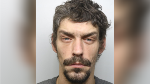 A custody photo of a man with dark hair, a dark beard, and moustache