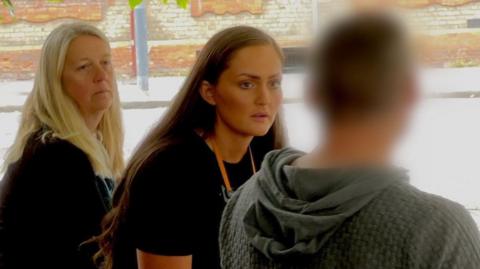 Ms Eddolls and Ms Davidson look at a man with concern. The man is blurred out in the image to preserve his identity. Both women are wearing black. The man is wearing a grey hoodie. They are sitting on a short stone wall in a square. 