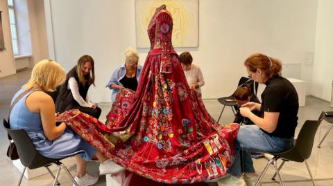 A red dress on a mannequin with women sat on chairs around the dress and embroidering it.
