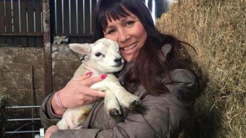 A woman in a brown coat is holding a lamb. She appears to be in an agricultural outbuilding and is stood next to bale of straw.