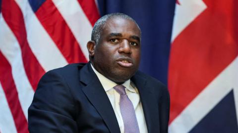 David Lammy looks ahead while sitting down in a dark blue suit and a patterned tie made up of small white and pink checks. The flags of the US and UK can be seen behind him.