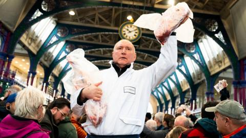 A meat trader in a white work jacket holds two joints of meat wrapped in white plastic. He is surrounded by customer, some of whom are holding up bank notes. The market clock is visible in the background of the image. 