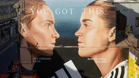 A mural painting on a side of the wall featuring the two football players, Jessica Fishlock and Alessia Russo. It shows their faces in profile, facing each other. Their hair is tied back and their football shirts are just visible at the bottom of the mural. 
