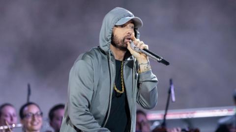 Eminem wearing a grey hoodie, golden chain against a black shirt, and grey pants performing on stage in front of an orchestra, as smoke fills the air around him