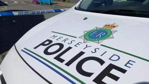 A white Merseyside Police vehicle seen parked close to some police tape, with the force's name and logo written on the bonnet. 