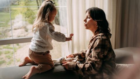 Mother and daughter spending time together at home, the mother is talking while the child plays on a sofa near a window