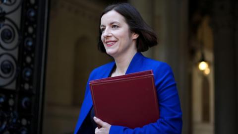 Bridget Phillipson smiling, wearing a blue suit and carrying a dark red folder