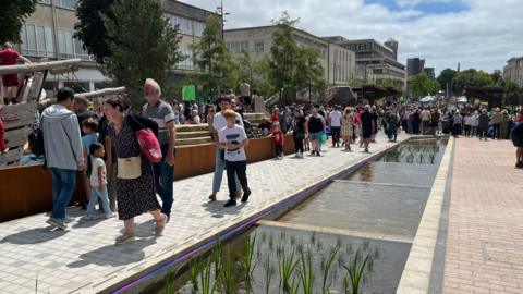 Members of people walking through a town centre alongside a long stretch of picturesque waterway which runs through the middle.