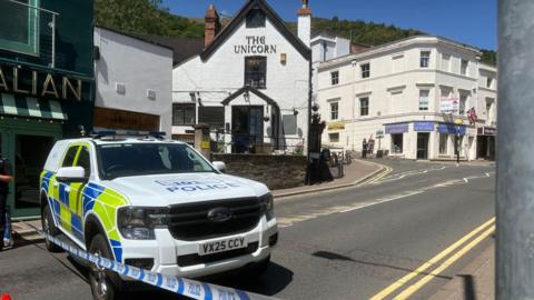 A police car and blue and white striped police tape in the foreground with an empty street behind and on the other side of the road a number of white buildings, one with the words The Unicorn on it