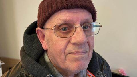 Alex Chapman, 74, is wearing a brown woollen bobble hat and has glasses and a dark overcoat on, with the hood down. The living room wall behind him is a magnolia colour.