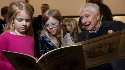 Skye (a girl with blonde hair and is wearing a pink top) she is stood next to Célestine (she is wearing a blue velvet top and has blonde hair and blue glasses) she is next to Nick Butterworth who is reading two books.